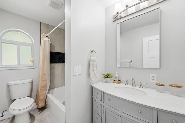 Bathroom featuring a built-in vanity with light gray cabinetry, an undermount sink, a large mirror, and a tiled shower with an arched window