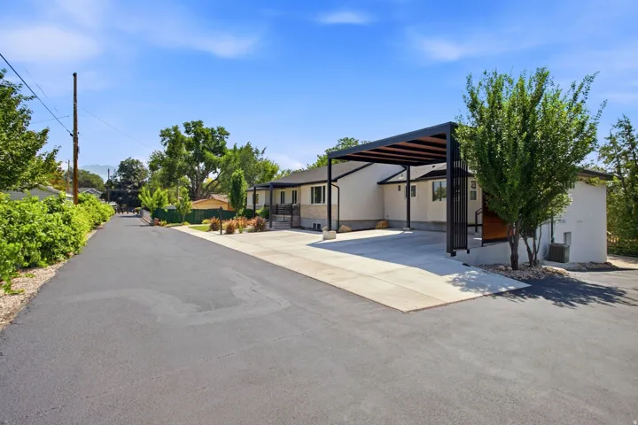 Paved driveway leading to a modern carport with slatted roof, stucco exterior, and mature landscaping