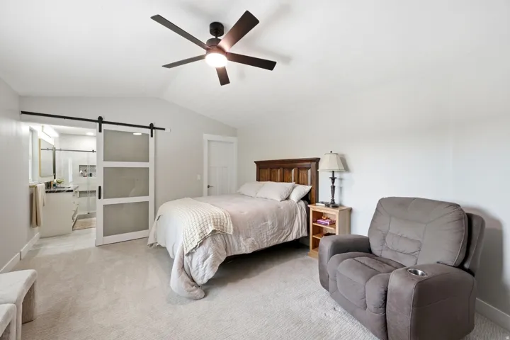 Spacious primary bedroom featuring neutral carpeting, wood-frame bed with headboard, ceiling fan with dark blades, and abundant natural light from multiple windows