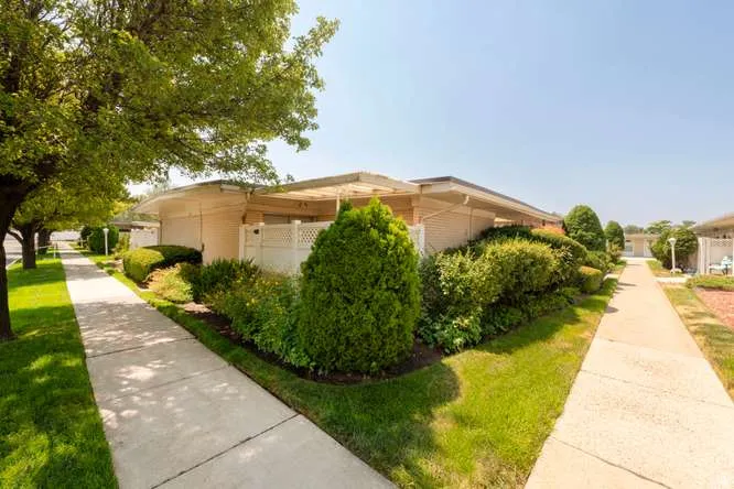 Charming single-story residence featuring a manicured landscape with mature trees and evergreen shrubs, concrete walkways, and lush lawn