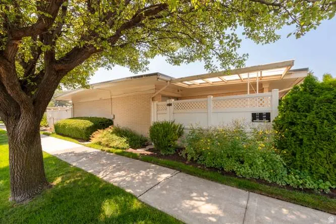 Cream brick exterior with an attached covered patio structure, concrete pathway, and mature landscaping