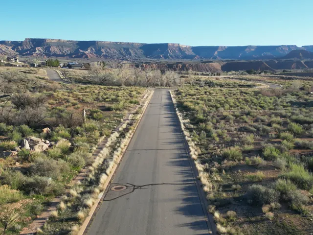 Paved road flanked by natural desert landscaping, featuring distant mesa formations and scattered residential structures