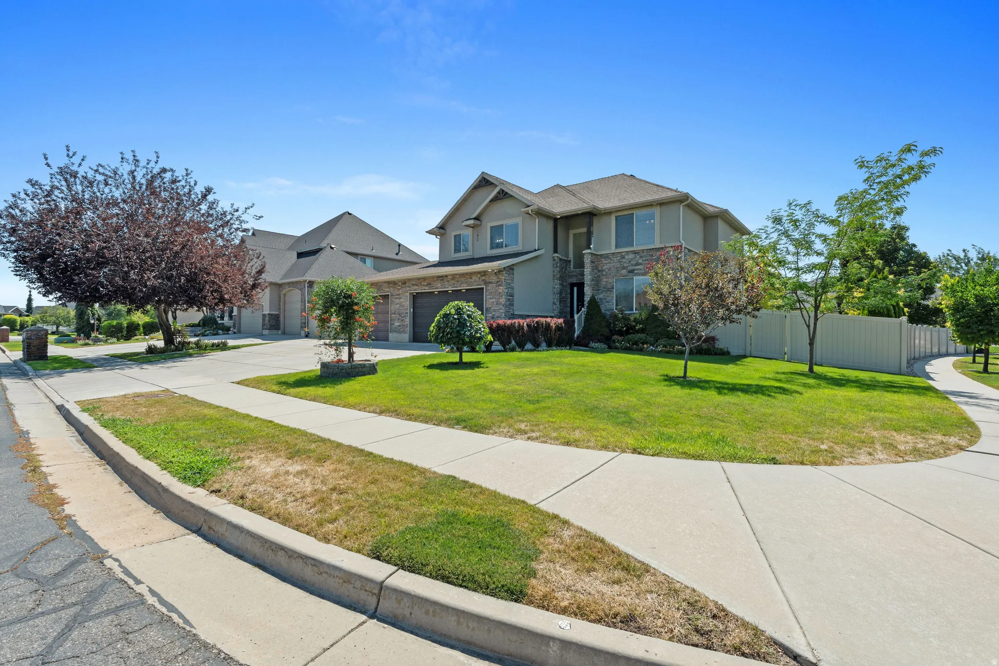 Stone and stucco exterior, multi-car garage, mature landscaping, concrete sidewalks, and a fenced yard