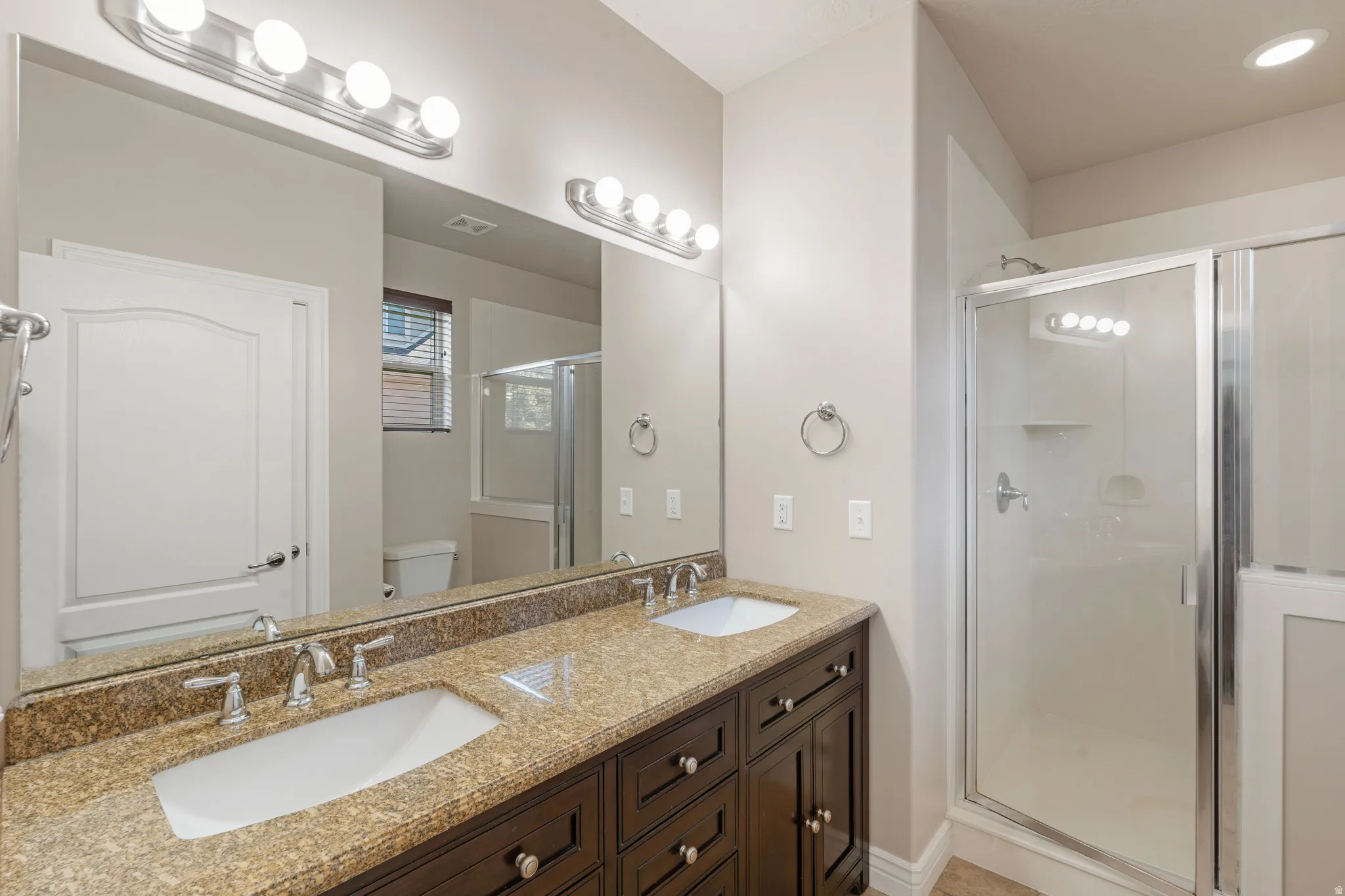 Dual vanity with integrated sinks, granite-finish countertop, dark wood-finish cabinetry, and a glass-enclosed shower