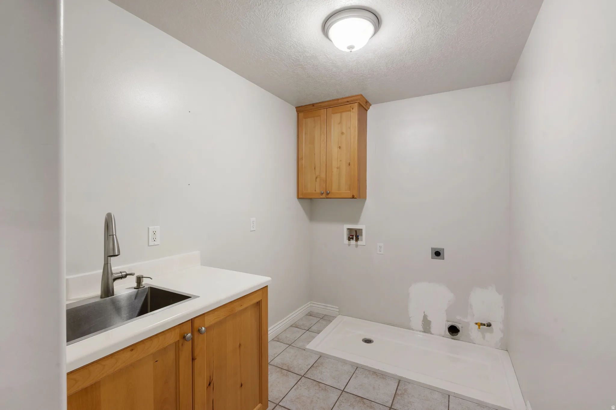 Laundry room featuring light wood-finish cabinetry, a stainless steel utility sink with a gooseneck faucet, light-toned countertop, and tiled flooring