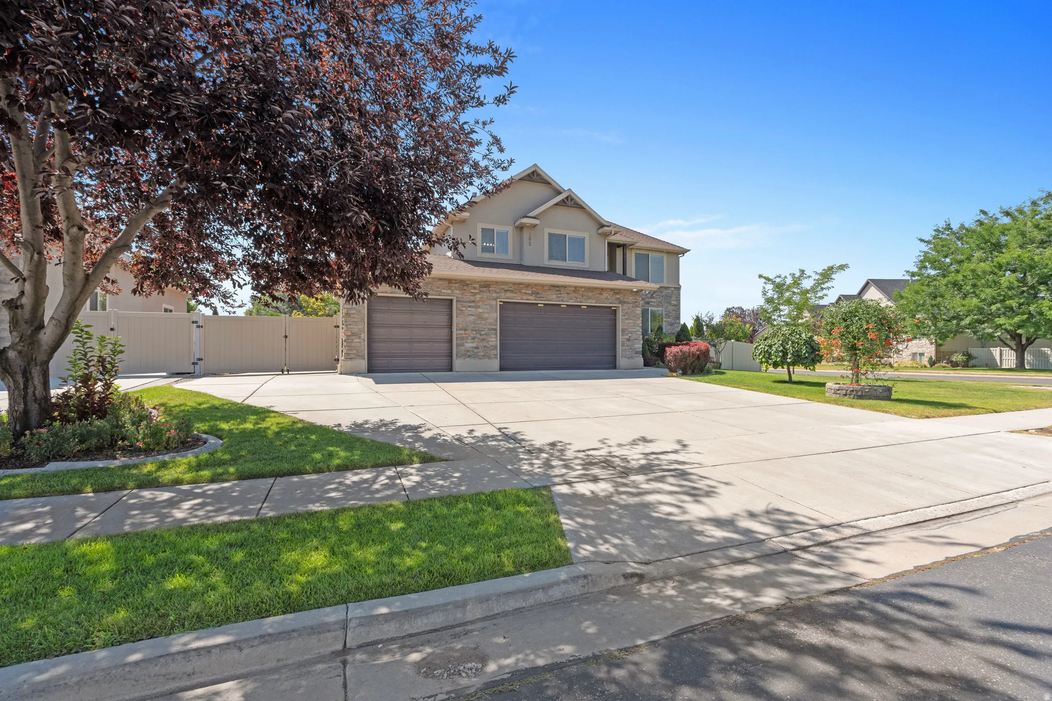 Two-story residence featuring a stone and stucco exterior, two attached garage doors, a concrete driveway, and front landscaping with mature trees and shrubs