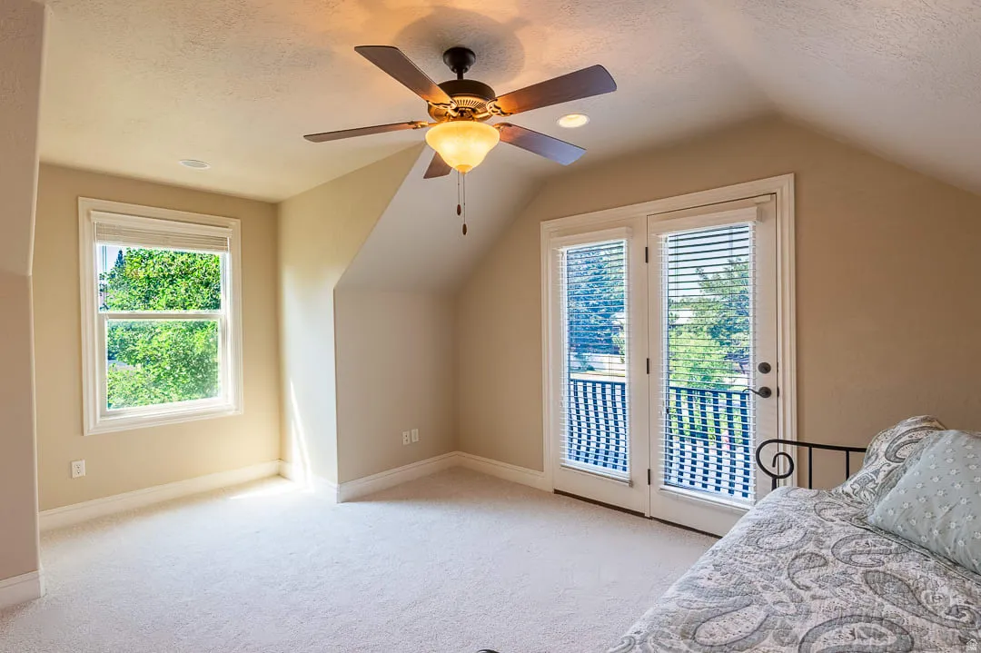 Carpeted flooring, vaulted ceiling, ceiling fan with light fixture, white framed windows, and a double-door balcony entrance