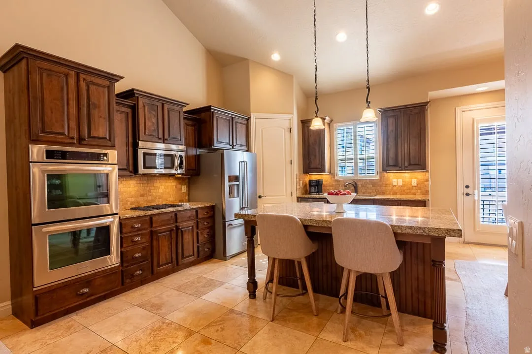 Kitchen featuring warm wood cabinetry, stainless steel appliances, a multi-tone tile backsplash, and an island with a stone countertop