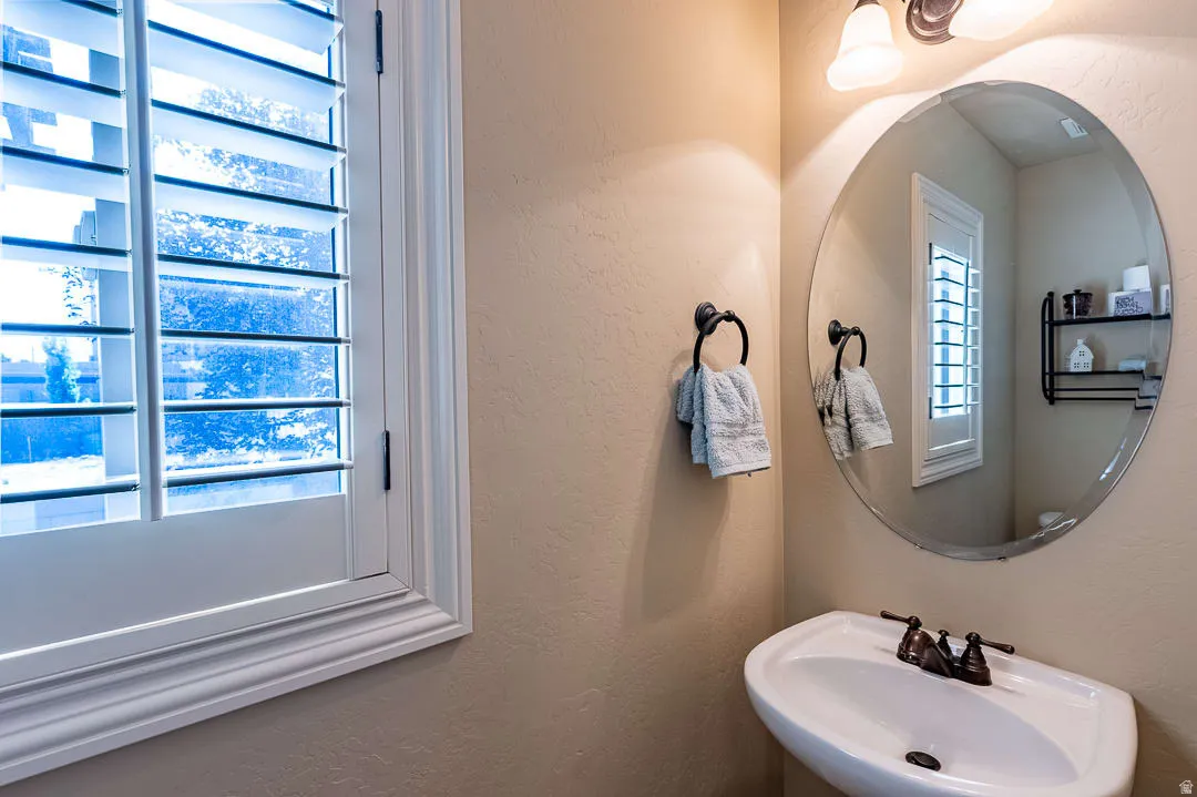 Window with white plantation shutters, oval mirror, wall-mounted light fixture, and pedestal sink with bronze-finish faucet