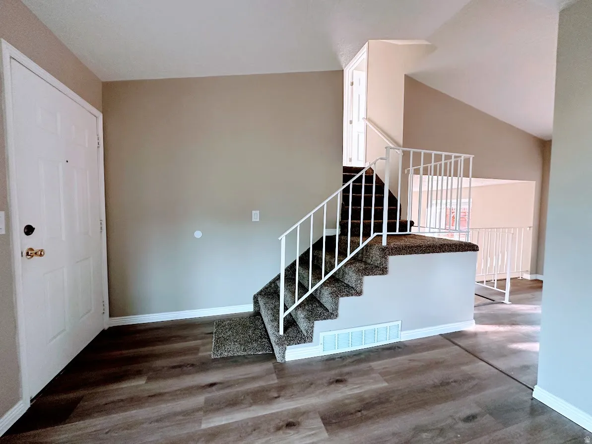 Wood-finish flooring, paneled entry door, carpeted staircase with white railing, and vaulted ceiling