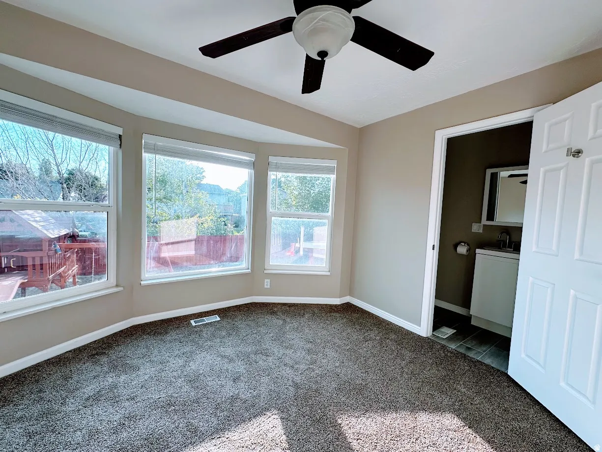 Bay window with multiple windows, ceiling fan, carpeted flooring, and an adjoining bathroom with a vanity, sink, and mirror