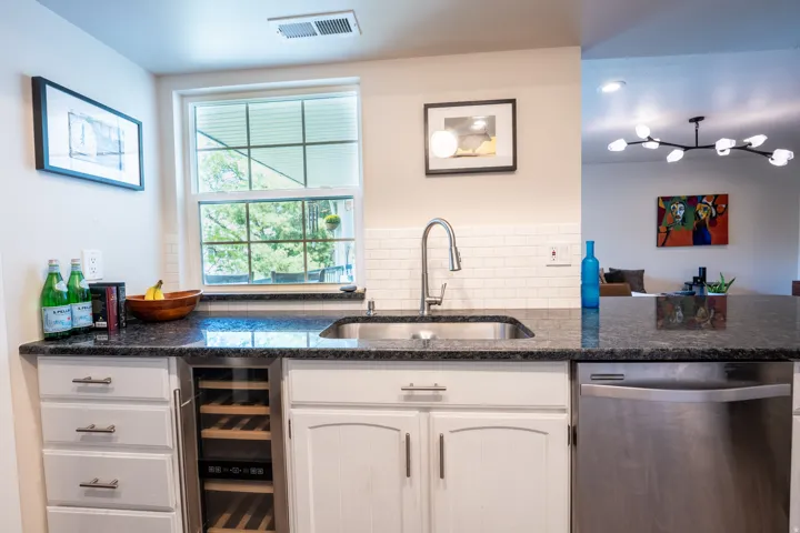 Stainless steel sink with a gooseneck faucet, white subway tile backsplash, dark stone countertops, integrated wine cooler, and stainless steel dishwasher