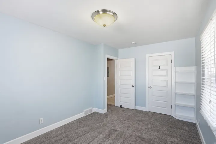 Plush carpet flooring, light blue wall paint, white trim, window with blinds, and a built-in shelving unit