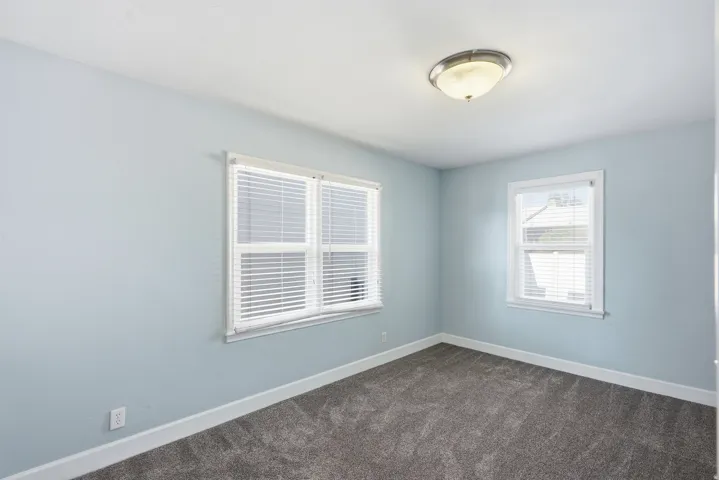 Bright bedroom with light blue walls, dual windows with white shutters providing natural illumination, soft-close ceiling fixture, and neutral carpet flooring throughout