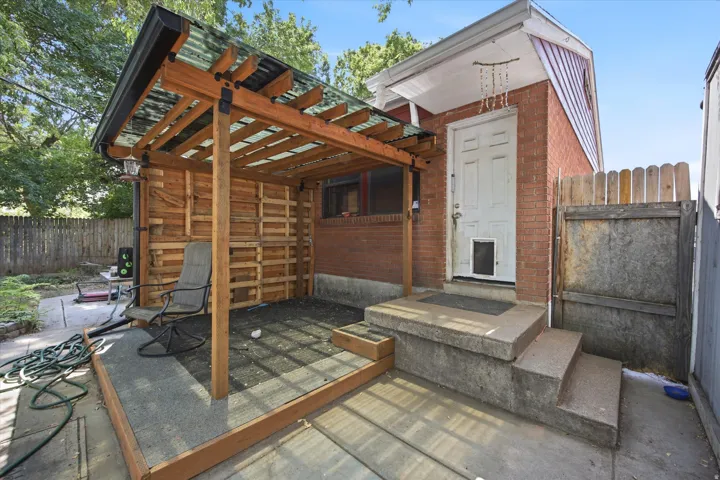 Brick exterior with a white entry door, concrete steps, and a sheltered patio featuring a wood-frame roof with translucent panels