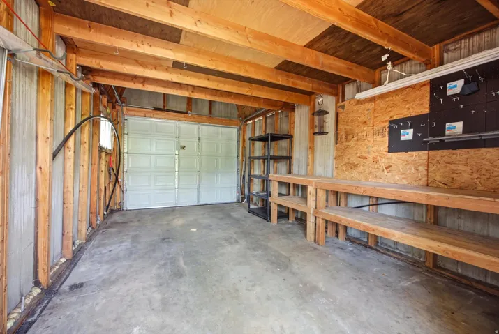 Concrete flooring, exposed wood beam ceiling, white paneled garage door, built-in shelving, and workbench