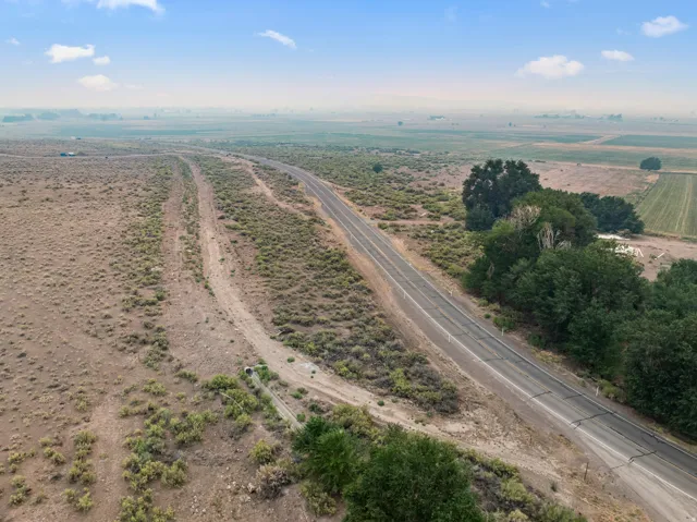 Paved road frontage alongside extensive, undeveloped land, featuring scattered green foliage and distant agricultural fields