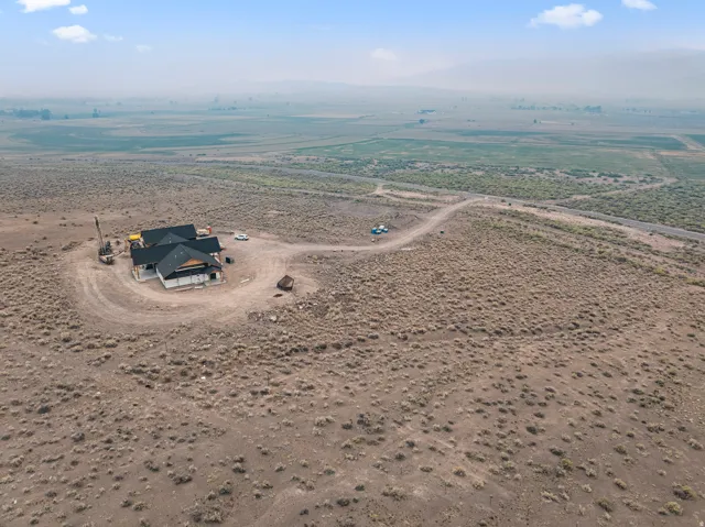 Newly constructed residence with a dark shingle roof, surrounded by expansive undeveloped land, featuring a winding dirt road entry