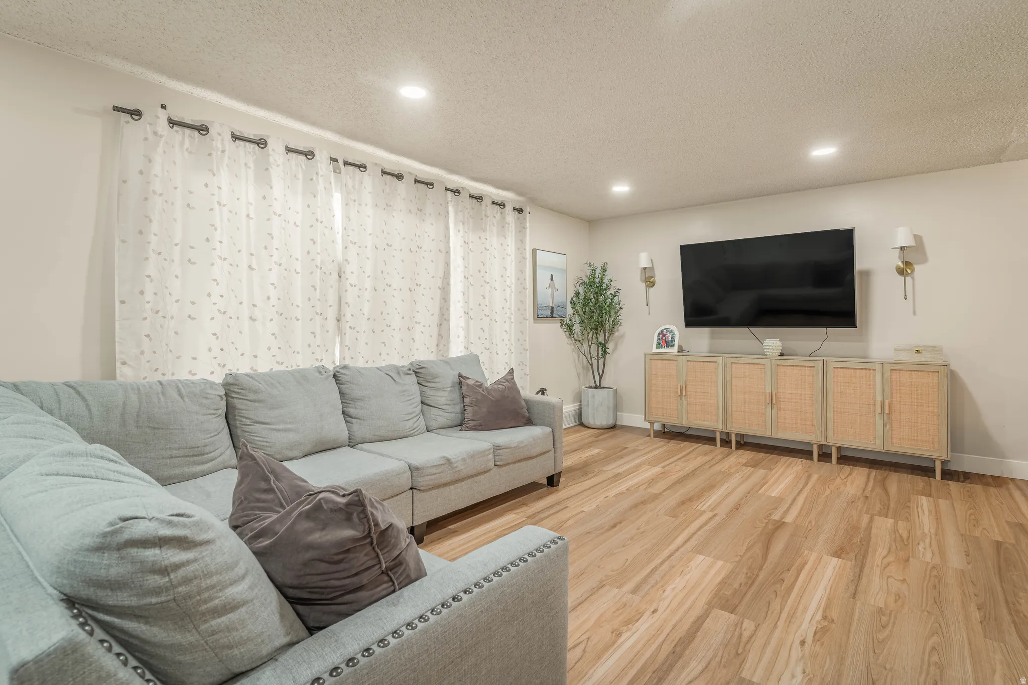 Bright living room showcasing warm wood-finish flooring, a spacious gray upholstered sectional sofa, and abundant natural light from large windows with sheer curtains