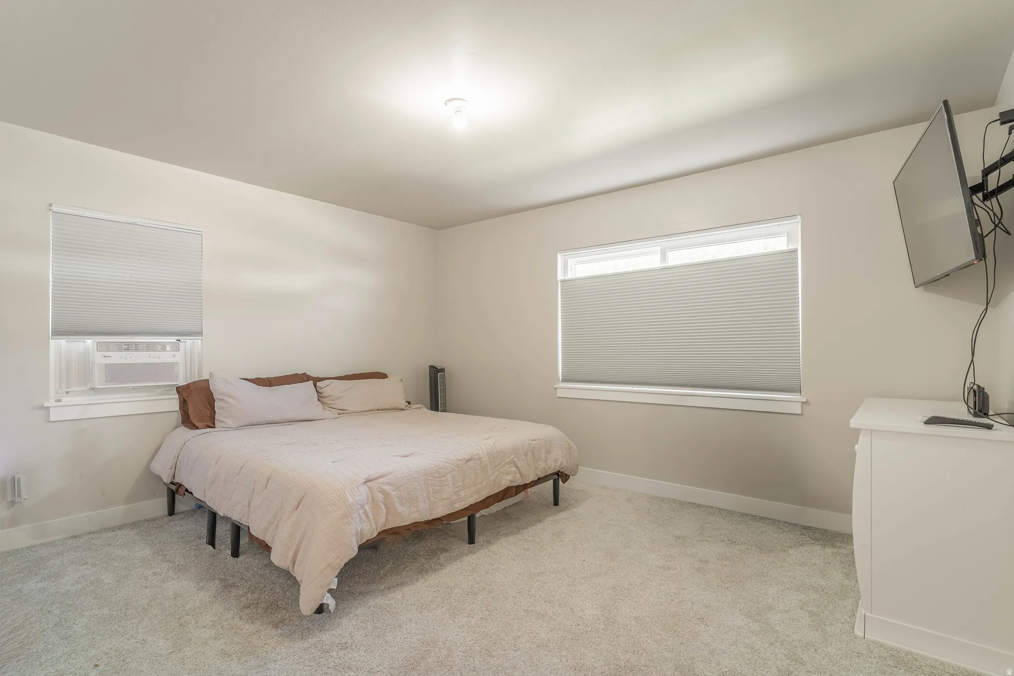 Spacious primary bedroom featuring neutral carpeting, a platform bed frame, dual windows with cellular shades allowing natural light, ceiling-mounted lighting, wall-mounted flat-screen television, and a white dresser with ample storage