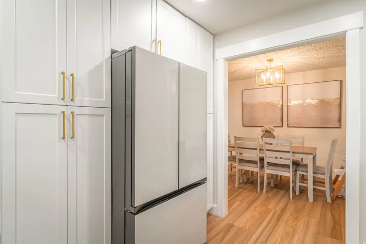 Modern kitchen featuring white shaker-style cabinetry with brushed gold hardware, stainless steel French-door refrigerator with bottom freezer, wood-finish flooring, and a seamless sightline into the adjacent dining area with pendant lighting and framed a