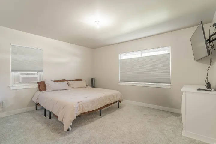 Spacious primary bedroom featuring neutral carpeting, a platform bed frame, dual windows with cellular shades allowing natural light, ceiling-mounted lighting, wall-mounted flat-screen television, and a white dresser with ample storage