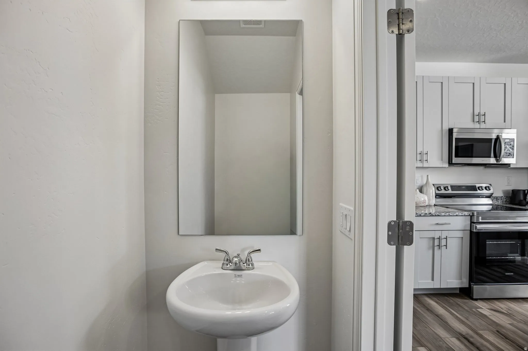 Pedestal sink with chrome faucet, framed mirror, light gray wall finish, wood-finish flooring, and light gray cabinetry with stainless steel appliances