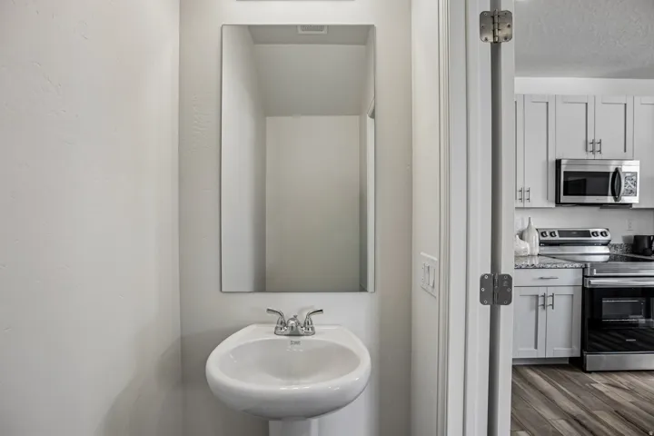 Pedestal sink with chrome faucet, framed mirror, light gray wall finish, wood-finish flooring, and light gray cabinetry with stainless steel appliances