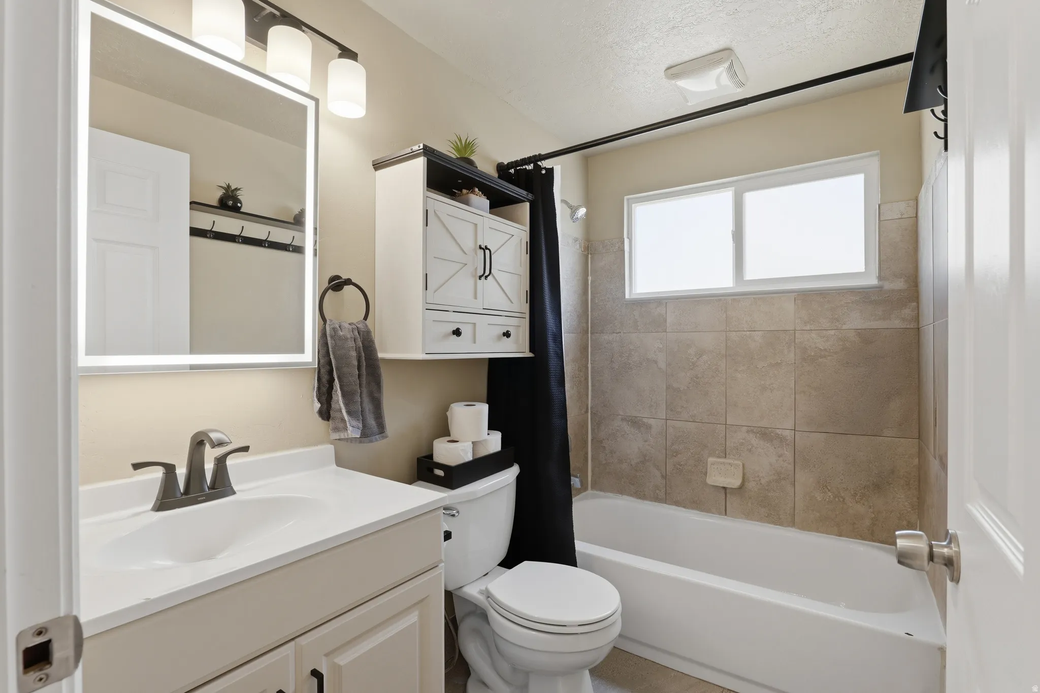 Vanity with integrated sink, backlit mirror, wall-mounted light fixture, and shower/tub combination with tiled surround