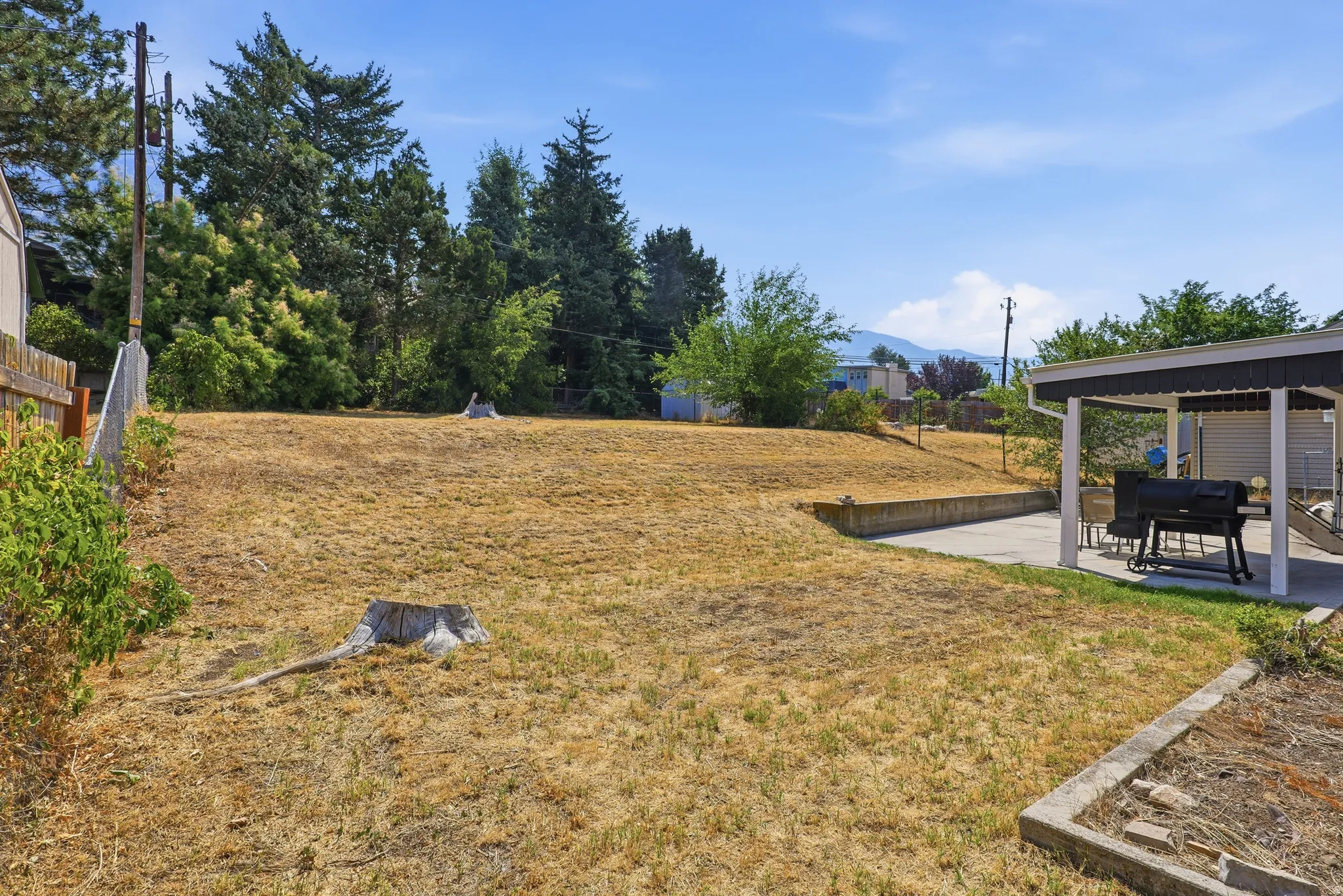 Expansive outdoor area featuring a covered patio, concrete hardscaping, natural terrain, and mature trees