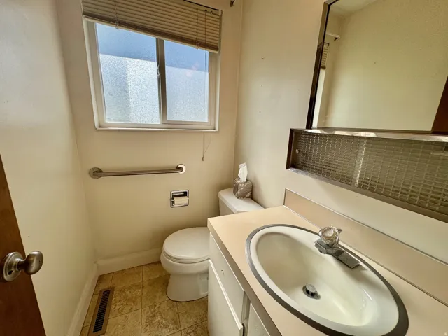Frosted window with horizontal blinds, vanity with integrated sink, stainless steel grab bar, and wall-mounted mirror with integrated lighting