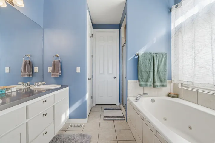Dual vanity with white cabinetry, built-in soaking tub, tiled flooring, and chrome fixtures