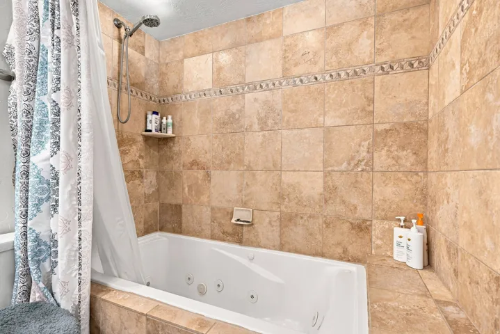 Built-in jetted tub surrounded by large format tile, decorative tile accent band, and a wall-mounted shower head with handheld attachment