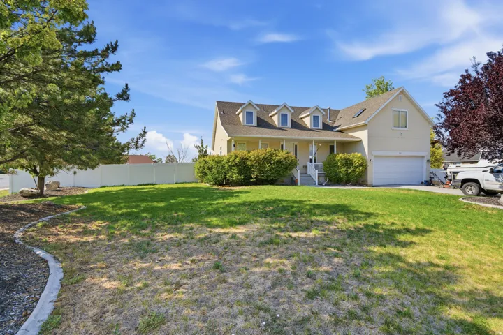 Gable roofline with three dormer windows, covered front porch, attached garage, and a lush front lawn with mature trees and shrubs