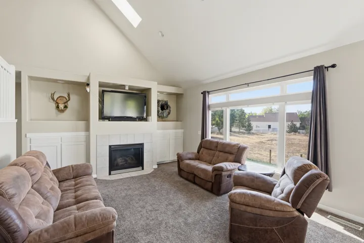 Vaulted ceiling with skylight, built-in shelving, tiled fireplace surround, large picture windows, and carpet flooring