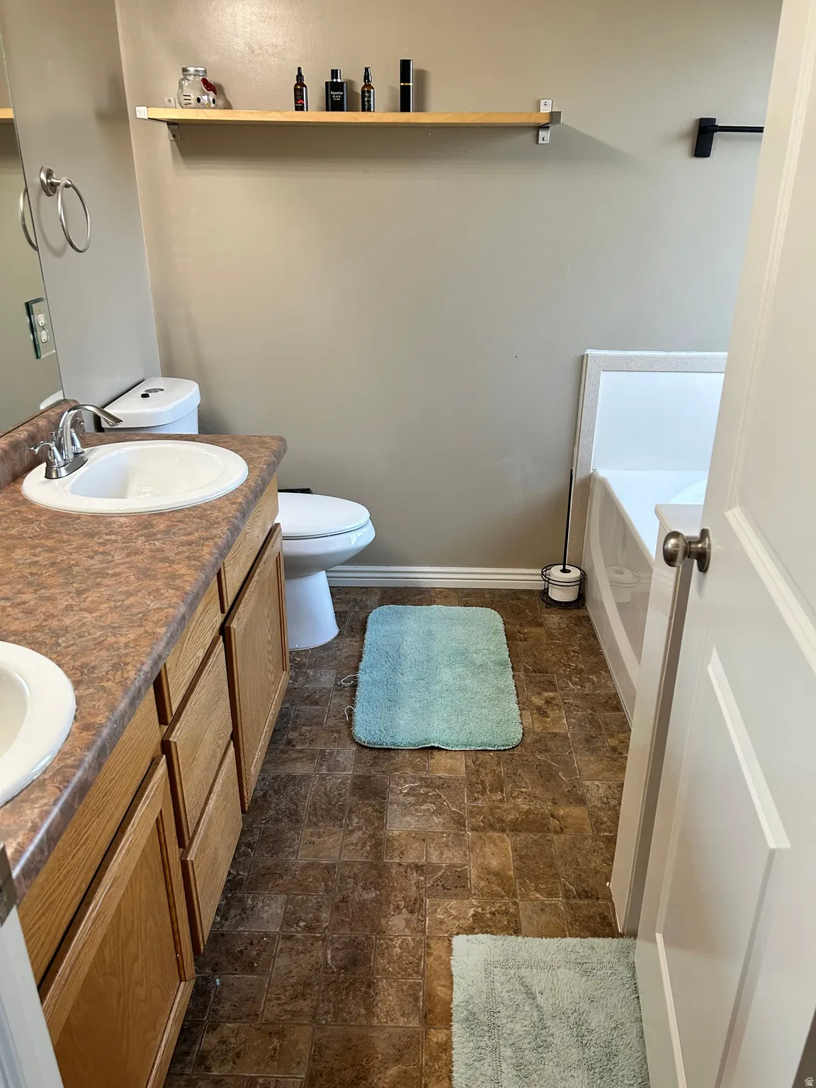 Dual-sink vanity with wood-finish cabinetry and stone countertops, ceramic tile flooring throughout, and coordinating bath accessories including toilet and towel bars
