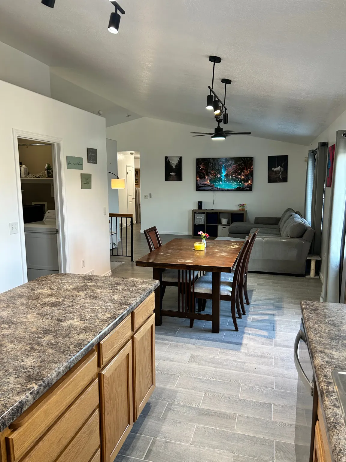 Vaulted ceiling with track lighting and ceiling fan, wood-finish kitchen cabinetry, speckled countertops, and plank-style flooring