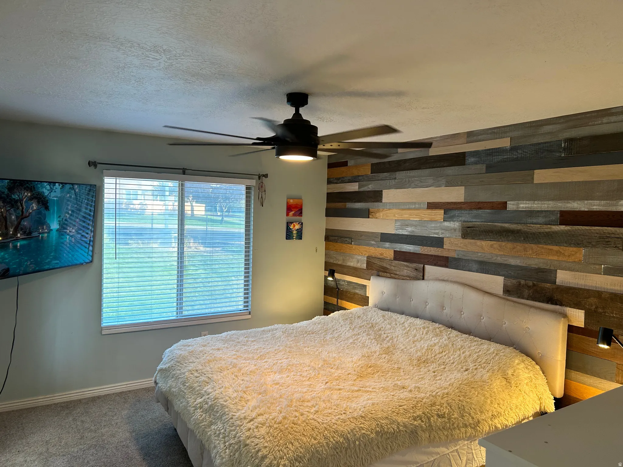 Textured accent wall, ceiling fan with integrated lighting, window with horizontal blinds, wall-mounted light fixtures, and carpet flooring