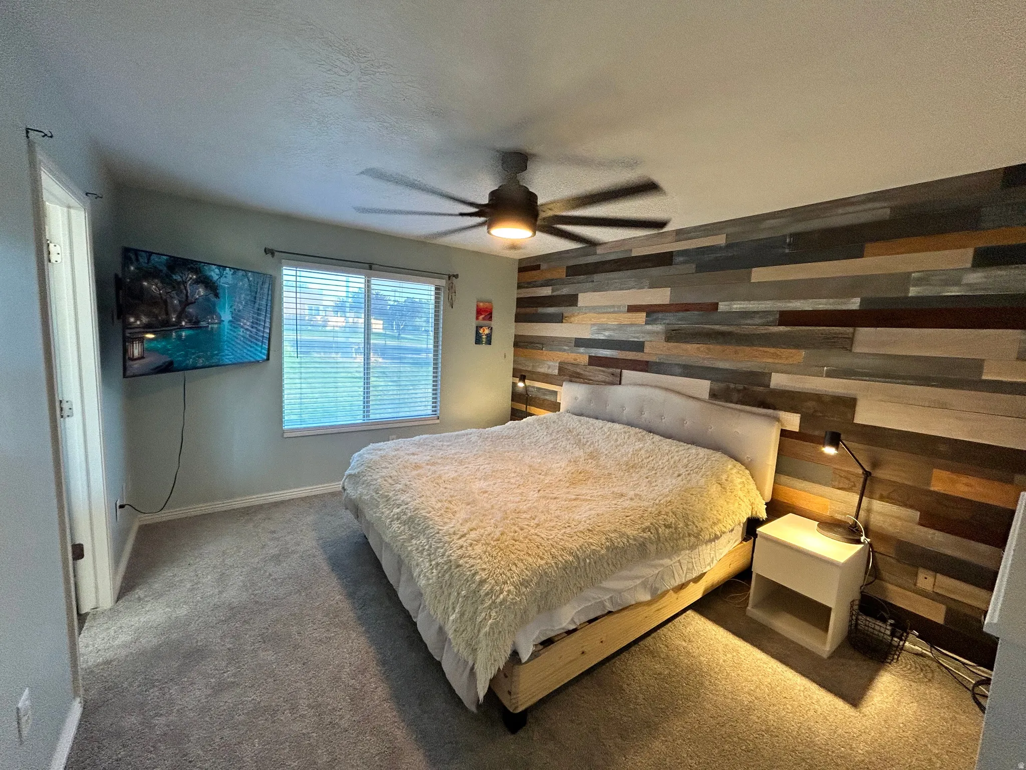 Wood-accented wall, window with blinds, ceiling fan with integrated lighting, carpeted flooring, and a wall-mounted television