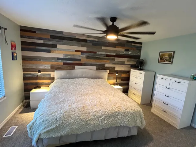 Accent wall featuring varied wood-finish planks, ceiling fan with integrated lighting, and neutral carpeting