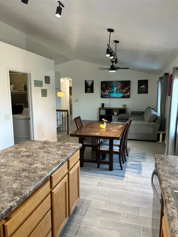 Vaulted ceiling with track lighting and ceiling fan, wood-finish kitchen cabinetry, speckled countertops, and plank-style flooring