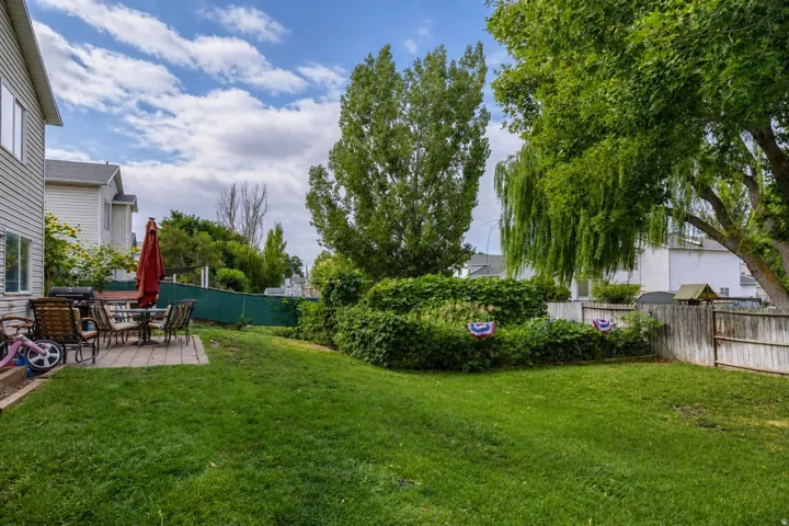 Expansive grassy yard featuring a paver patio, a partial privacy fence, mature trees, and established perimeter foliage