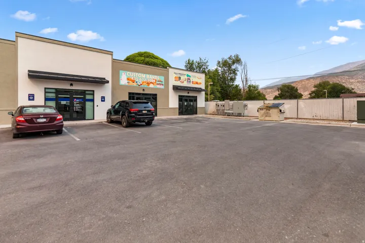 Paved parking lot with multiple spaces, commercial storefronts with large glass entry doors, and a mountain backdrop