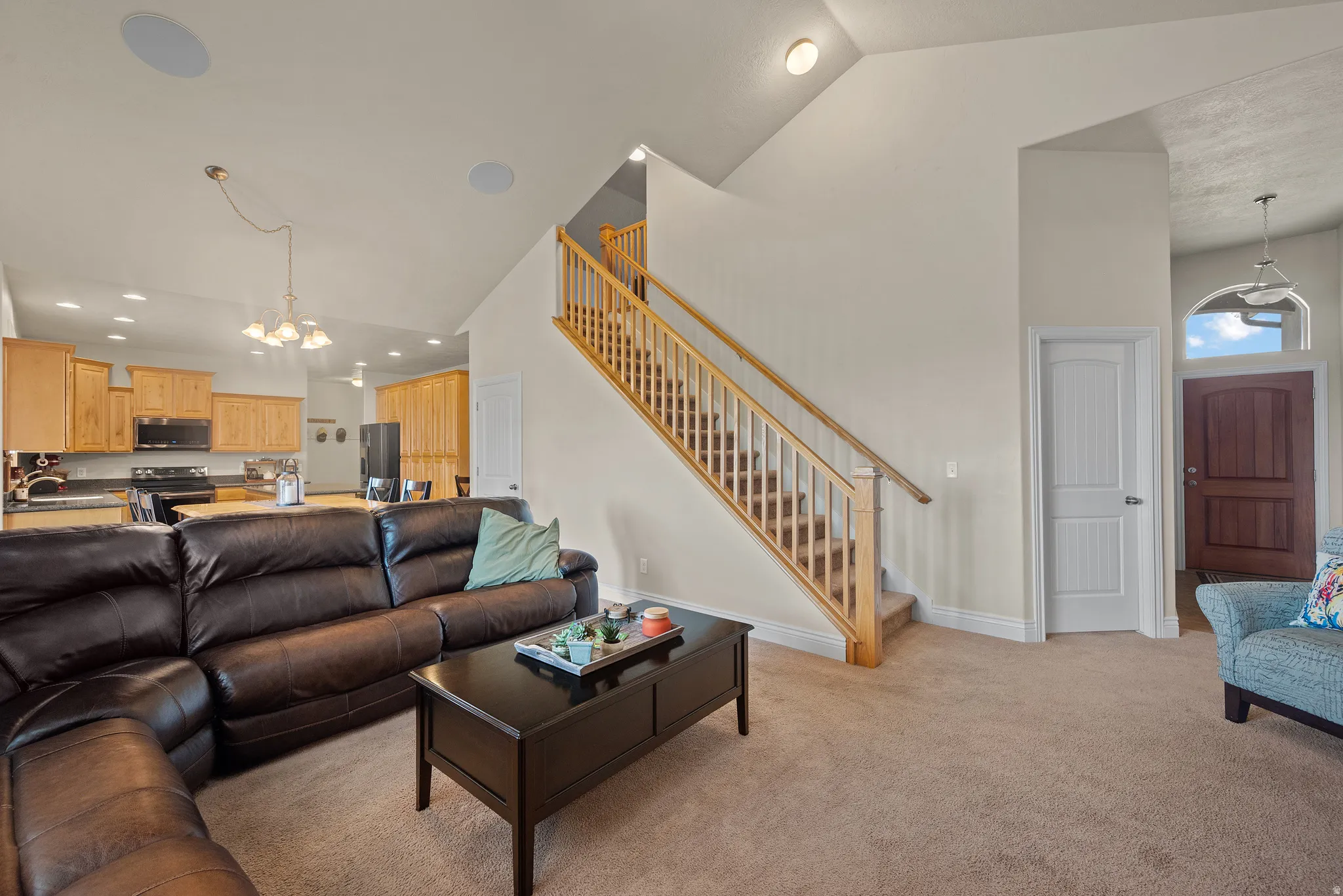 Open-concept living space featuring vaulted ceilings, a prominent staircase with wood railing, warm wood-finish kitchen cabinetry, and an arched entry door with transom window