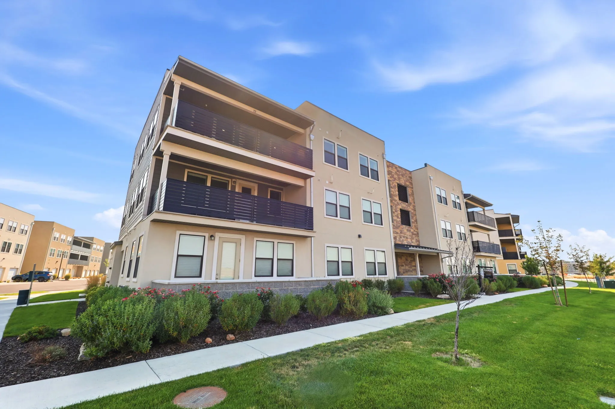 Multi-story structure featuring a stucco exterior, stone accent cladding, multiple balconies with horizontal railings, and ground-level landscaping