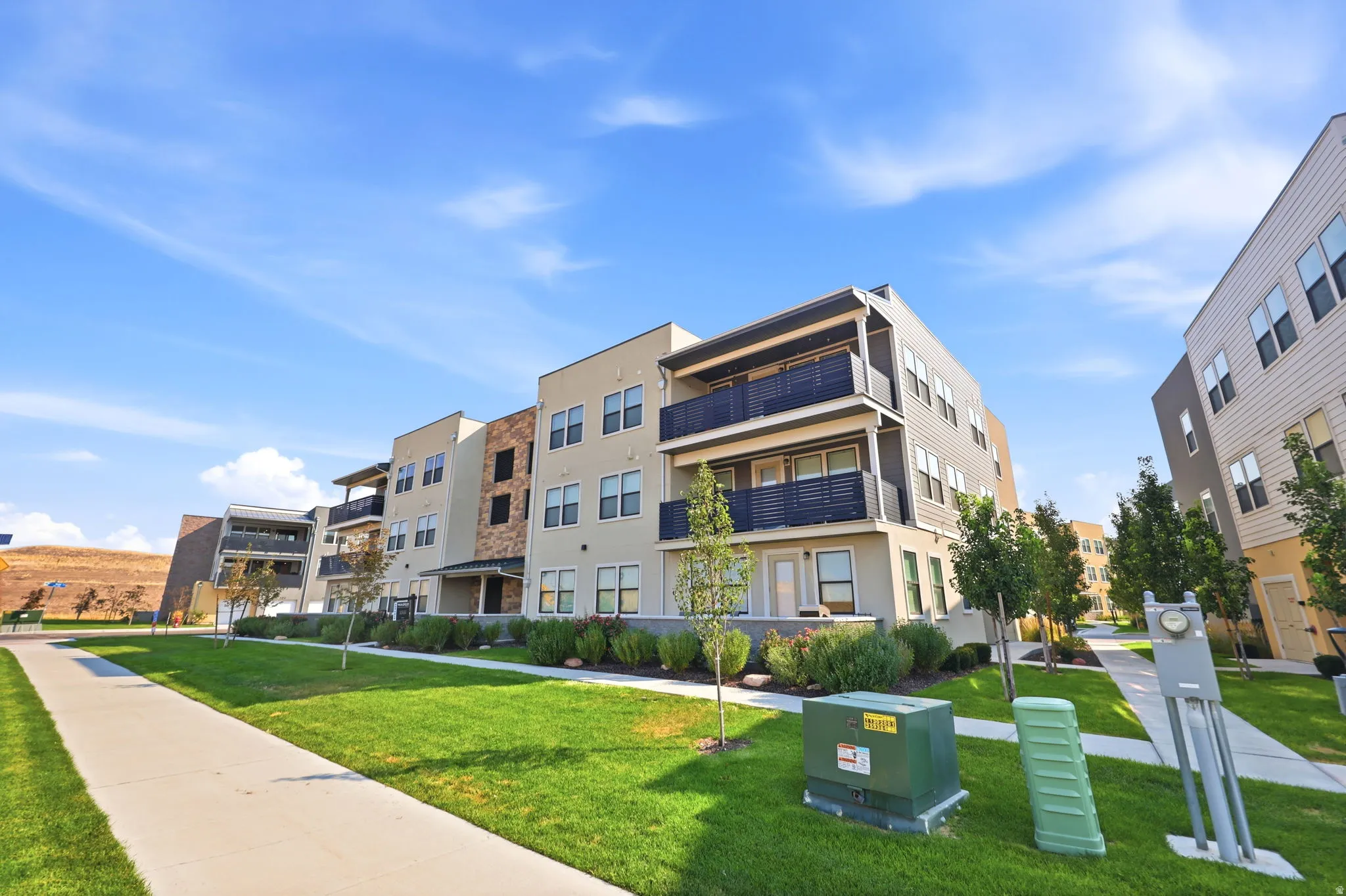 Contemporary multi-story building featuring multiple balconies, a mix of siding and stone veneer exterior, ground-level patio, and surrounding green space