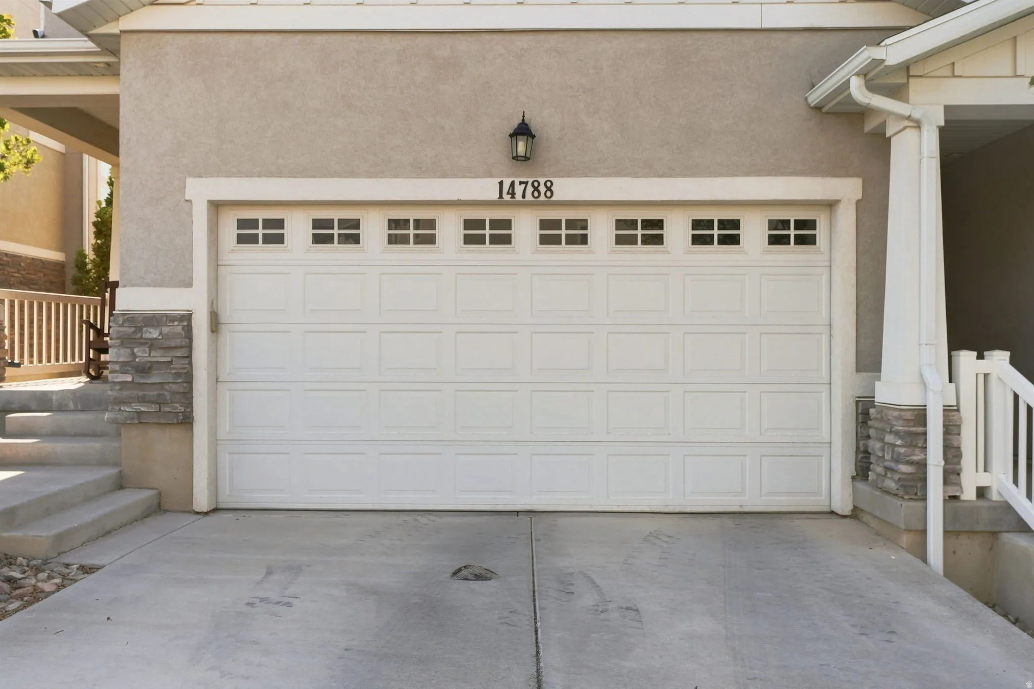 White paneled garage door with upper window inserts, stucco exterior, exterior wall lantern, stone veneer accents, and concrete driveway
