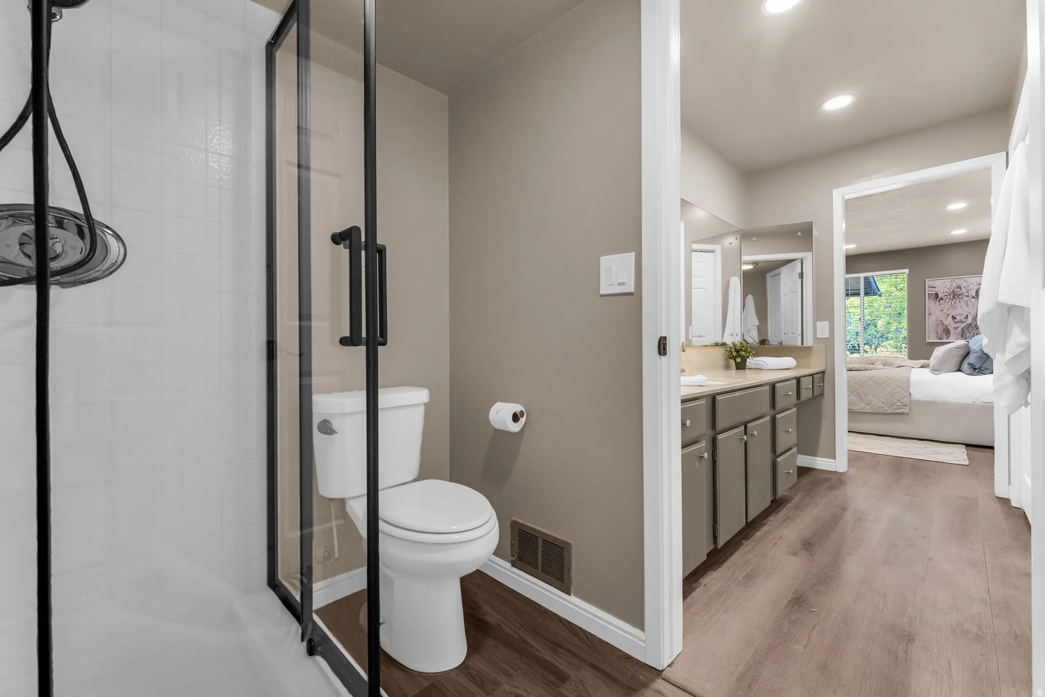 Glass-enclosed shower with a matte black frame, white tiled shower surround, dual vanity with undermount sinks, and wood-finish flooring