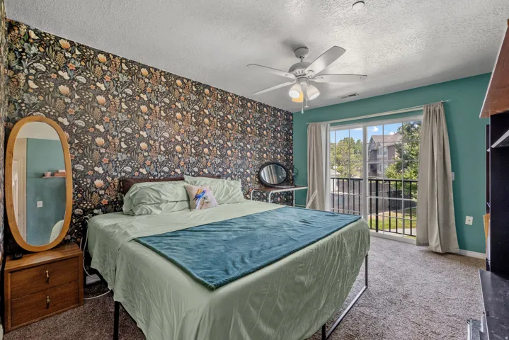 Textured wall finish, ceiling fan with integrated lighting, sliding glass door, private balcony with black metal railing, and carpeted flooring