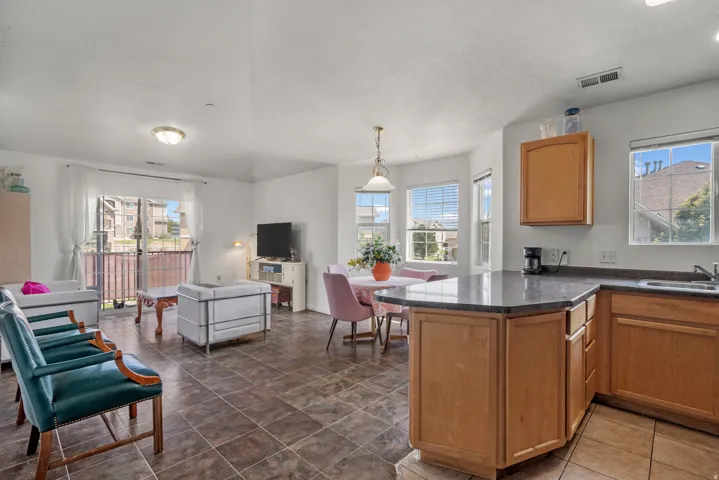 Open-concept living space with tiled flooring, a kitchen featuring wood-finish cabinetry and stone-finish countertops, a dedicated dining area with pendant lighting, and abundant natural light from multiple windows and a sliding glass door